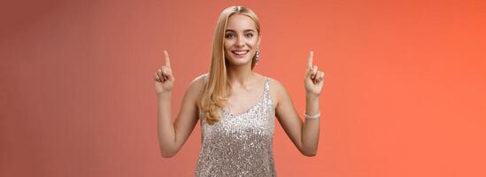 Charming feminine tender blond woman in silver party dress raise hands pointing up smiling delighted recommend awesome cosmetics good product service, standing happily grinning red background photo