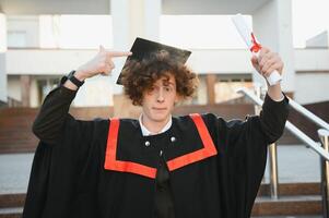 Happy smiling university graduate in mantle holding diploma in raised hand and expressing happiness over university building at background. Successful graduating from university or college photo