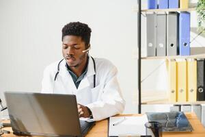 Smiling african american female doctor gp wears white medical coat using laptop computer at workplace gives remote online consultation, working on pc, consulting patient in internet telemedicine chat photo