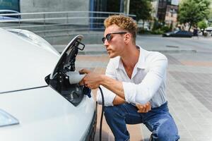 Stylish man inserts plug of the charger into the socket of electric car close-up photo