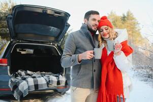 Couple in love sitting in car trunk drinking hot tea in snowy winter forest and chatting. People relaxing outdoors during road trip. Valentines day photo
