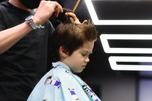 Side view of cute little boy getting haircut by hairdresser at the barbershop. photo