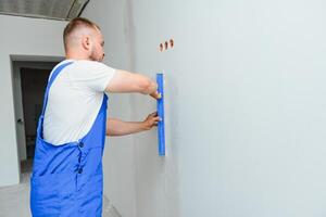 The builder carefully corrects the irregularities of the wall with a trowel. Builder in work clothes against a gray wall. Photo plasterer at work.