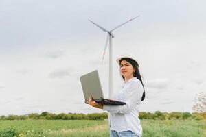 cerca arriba retrato de hembra ingeniero en casco en pie y utilizando ordenador portátil computadora mientras comprobación el trabajo de molino turbina a renovable energía estación. foto