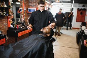 un Barbero es yendo mediante el eléctrico corte y afeitado máquina para el barba de un afroamericano brasileño chico. foto