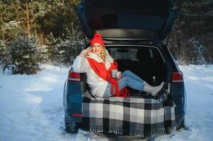 young woman sitting in car trunk drinking warm tea and takes a selfie at winter snowed day. photo