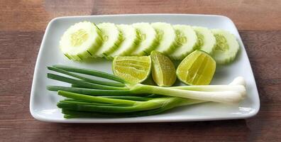 Fresh Scallion, sliced cucumber and lime or lemon in white plate on wooden table. Organic vegetable food and Harvest of agriculture concept photo