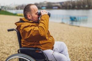 parapléjico minusválido hombre en silla de ruedas es utilizando prismáticos exterior. él es acecho naturaleza por el lago. foto