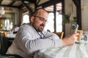 contento parapléjico minusválido hombre en silla de ruedas es sentado a restaurante y utilizando teléfono inteligente foto