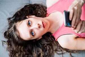 Young woman with long curly hair lies on the sofa and scrolls social networks on her phone in a pink top, top view photo