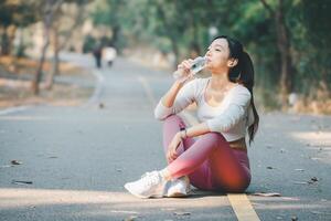 Fitness concept, An athletic woman sitting on a park road takes a water break after exercising, enjoying her workout routine. photo