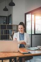 Business woman concept, A focused businesswoman takes notes in a notepad while working on her laptop in a well-lit workspace with modern decor. photo