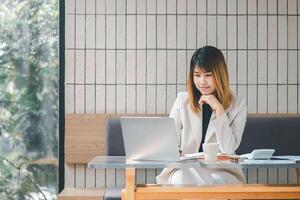 A pensive businesswoman in deep thought while working on her laptop at a well-lit cafe, surrounded by her work materials. photo