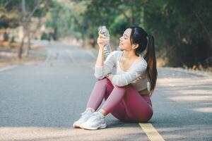 Sport fitness concept, A cheerful young woman sitting on a forest road takes a water break after a run, enjoying her fitness routine outdoors. photo