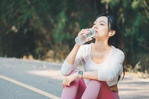 Fitness concept, An athletic woman sitting on a park road takes a water break after exercising, enjoying her workout routine. photo