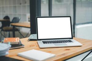 An open laptop with a blank screen sits on a wooden desk, surrounded by notebooks and a pen, in a bright, modern workspace. photo