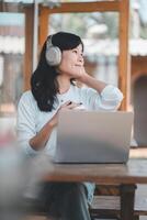 Business freelance concept,A contented woman with headphones enjoys music as she works on her laptop at a well-lit table with a coffee and plant nearby. photo