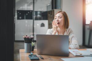 Exhausted female office worker yawning in front of her laptop during a busy workday, indicating long hours or lack of sleep. photo