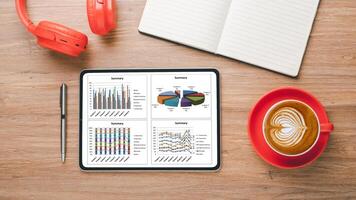 Top view of a digital tablet displaying colorful business analytics graphs, accompanied by a notebook, pen, coffee, and headphones on a wooden table. photo