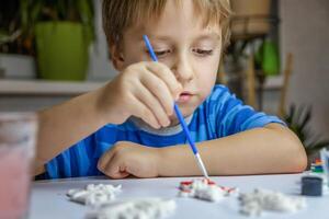 The kid draws and paints plaster figures with a brush and gouache on a piece of paper. photo