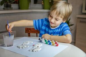 The kid draws and paints plaster figures with a brush and gouache on a piece of paper. photo