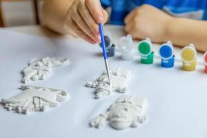 The kid draws and paints plaster figures with a brush and gouache on a piece of paper. photo