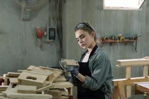 A young woman is training to be a carpenter in the workshop. She works with a laptop computer in a wood workshop. female carpenter contact customers by smartphone. SME orders, Start-ups and small photo