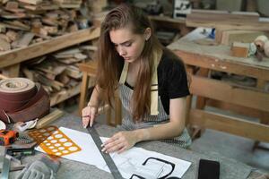 A young woman is training to be a carpenter in the workshop. She works with a laptop computer in a wood workshop. female carpenter contact customers by smartphone. SME orders, Start-ups and small photo