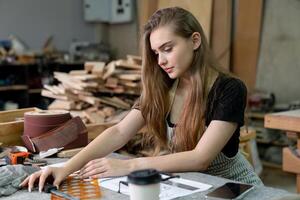 A young woman is training to be a carpenter in the workshop. She works with a laptop computer in a wood workshop. female carpenter contact customers by smartphone. SME orders, Start-ups and small photo