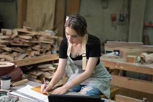 un joven mujer es formación a ser un carpintero en el taller. ella trabajos con un ordenador portátil computadora en un madera taller. hembra carpintero contacto clientes por teléfono inteligente SME foto