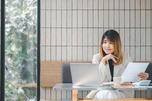 A confident businesswoman assesses documents with a pen in hand, laptop open, in the bright ambiance of a cafe setting. photo