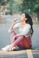 Fitness concept, An athletic woman sitting on a park road takes a water break after exercising, enjoying her workout routine. photo