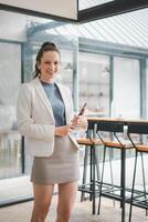 Business woman concept, A professional woman in a smart casual outfit stands with a phone in a contemporary cafe setting, exuding confidence and readiness. photo