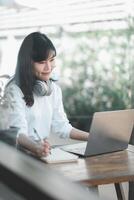 un alegre joven mujer con un computadora portátil, sentado a un de madera mesa en un acogedor cafetería, contemplando con un agradable sonrisa. foto