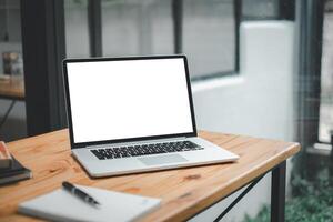 Open laptop with a white blank screen sitting on a wooden table in a bright, modern workspace with notebook and pen. photo