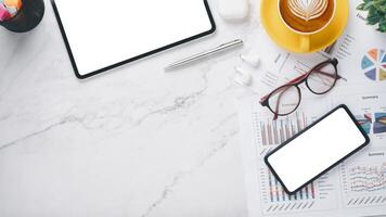 Modern work desk featuring two blank screen devices, coffee cup, eyeglasses, and business documents, set on a marble background. photo