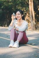 Sport fitness concept, A joyful young woman takes a water break outdoors after exercising, wearing earphones and sportswear, embodying a healthy lifestyle. photo