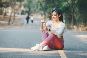 Sport fitness concept, A cheerful young woman sitting on a forest road takes a water break after a run, enjoying her fitness routine outdoors. photo