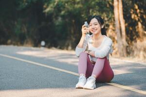 Sport fitness concept, A cheerful young woman sitting on a forest road takes a water break after a run, enjoying her fitness routine outdoors. photo