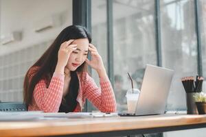 A stressed young woman holds her head in frustration while working at her laptop in a cluttered office environment. photo