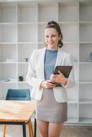 Business management concept, A smiling young professional woman stands in a modern office, holding a tablet, ready for a productive workday. photo