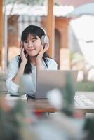 un contento joven asiático mujer con auriculares y un teléfono inteligente disfrutando música mientras trabajando en su ordenador portátil afuera. foto