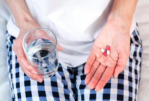 Young woman holds two pills and glass of water in hands. Taking vitamins, supplements, antibiotic, antidepressant, painkiller medication. Prenatal vitamins. Close-up. photo