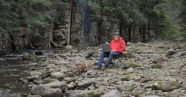 A man works on a laptop near a stream in nature. Concept of freelancing, digital nomad or remote office. 4K video