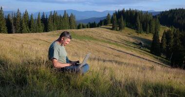 un' uomo si siede su un' bellissimo prato nel il montagne, lavori su un' il computer portatile. concetto di libero professionista, digitale nomade o a distanza ufficio. video