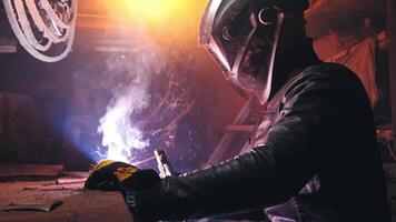 A welder in a protective mask and leather jacket is welding in his atmospheric workshop. Low key. Real time video