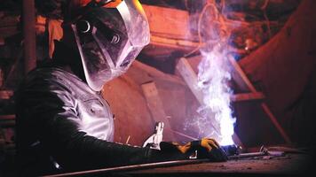 A welder in a protective mask and leather jacket is welding in his atmospheric workshop. Low key. Real time video