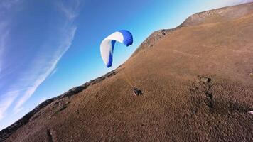 A paragliding athlete flies on a paraglider near the slope of a small mountain near a village on a sunny autumn day. Aerial view shooting with FPV drone quadcopter in acro mode video