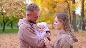 giovane madre e padre siamo a piedi con un' neonato bambino nel il autunno parco. genitori nel identico tute da ginnastica Sorridi e Guarda a il bambino video