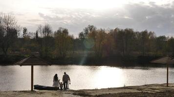 un familia disfruta un caminata cerca un lago. madre, padre y pequeño niño hija descansando a puesta de sol en primavera y celebrando mundo internacional día video